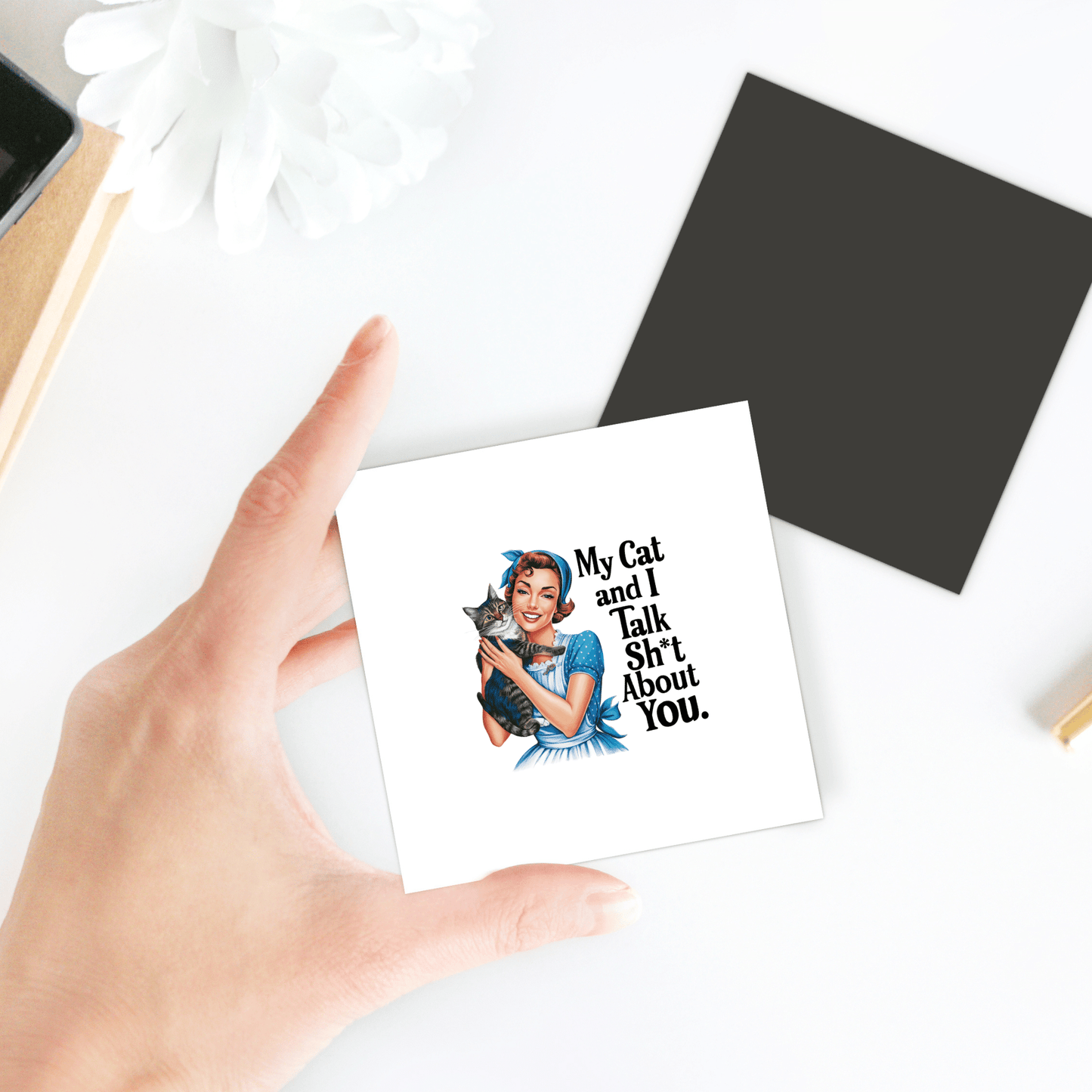 A hand holds the My Cat and I Talk Sh*t Magnet by Mirabilia Boutique, a 3x3 retro-style UV printed fridge magnet for cat lovers. A black square and white flower sit on the table in the background.