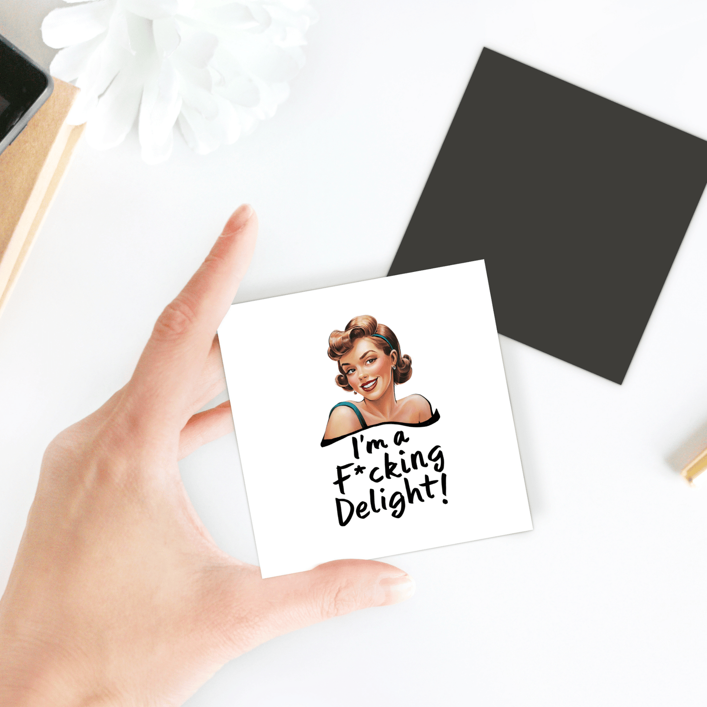 A hand holds the Mirabilia Boutique “I’m a F*cking Delight” 3x3 retro magnet—a funny, sassy gift—against a background with a black card, white flower, and desk items.