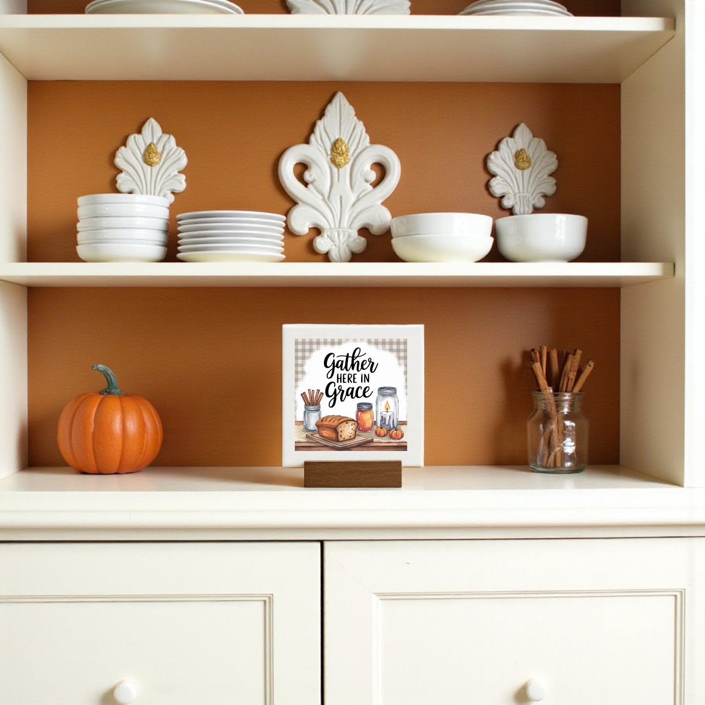 A white kitchen hutch displays stacked white dishes, bowls, farmhouse decor, a small pumpkin, a jar with cinnamon sticks, and the Mirabilia Boutique Mini Gather Here in Grace Sign (4x4), perfect for charming shelf decor.