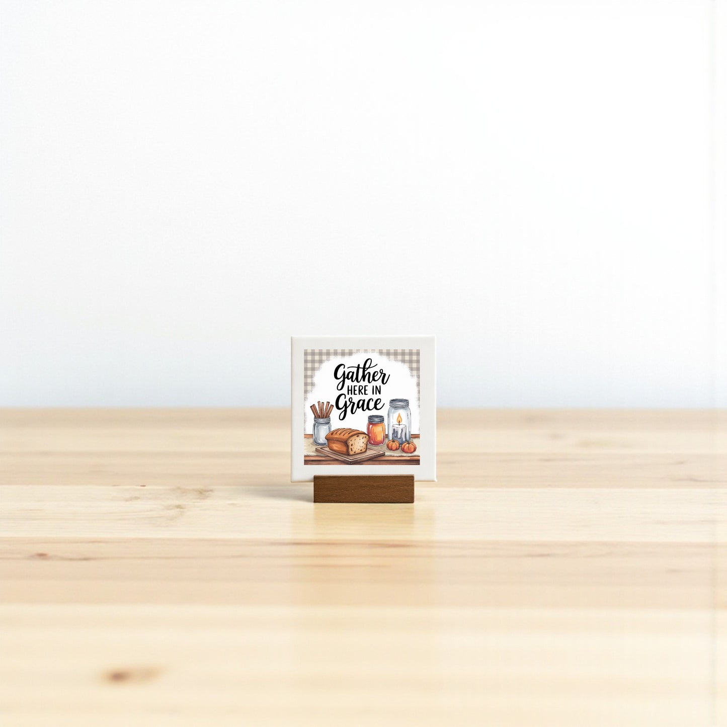 The Mirabilia Boutique Mini Gather Here in Grace Sign (4x4) sits on a wooden table with farmhouse accents—bread, a rolling pin, and kitchen decor—making it a charming Thanksgiving gift against a crisp white background.