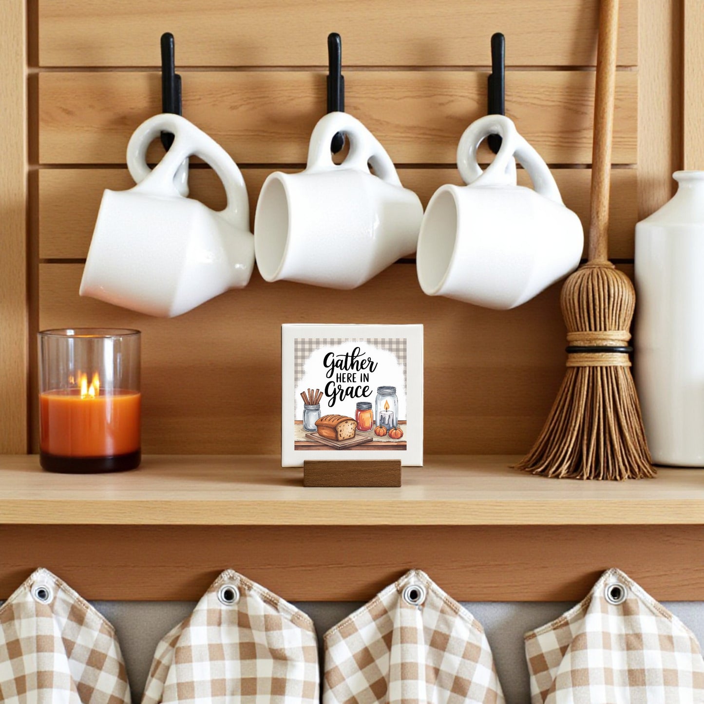 A cozy kitchen shelf displays a lit candle, three white mugs, a small broom, gingham fabric, and the Mirabilia Boutique Mini Gather Here in Grace Sign (4x4)—a charming farmhouse touch.