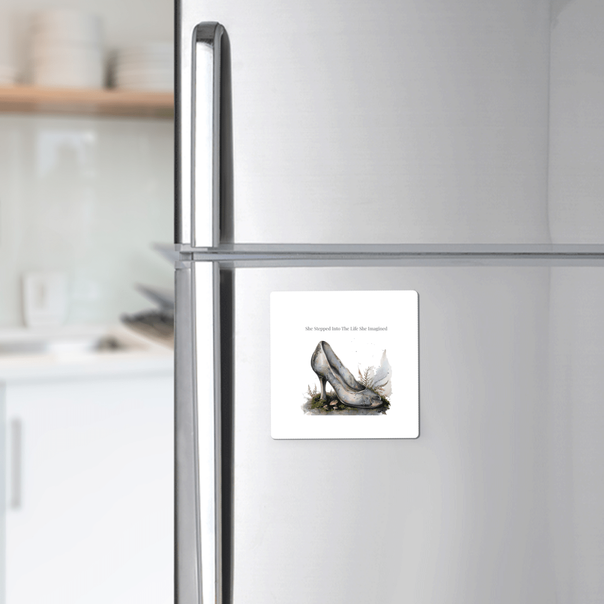 A silver refrigerator displays the Mirabilia Boutique "She Stepped Into the Life She Imagined Magnet" on its freezer door—a square, inspirational magnet illustrated with a glass slipper and uplifting text—while the softly blurred kitchen background enhances the enchanting, fairytale-inspired decor.