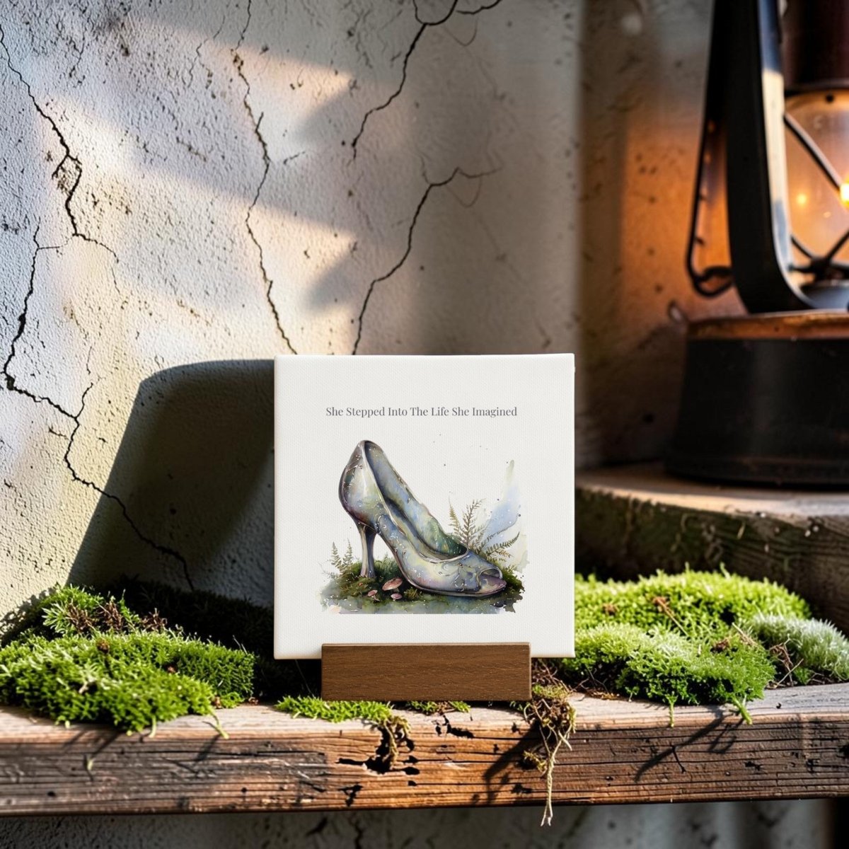 The "She Stepped Into the Life She Imagined" Mini Sign by Mirabilia Boutique features a painted high-heeled shoe, evoking glass slipper artistry, and is elegantly displayed on a wooden stand atop a mossy shelf beside a vintage lantern.