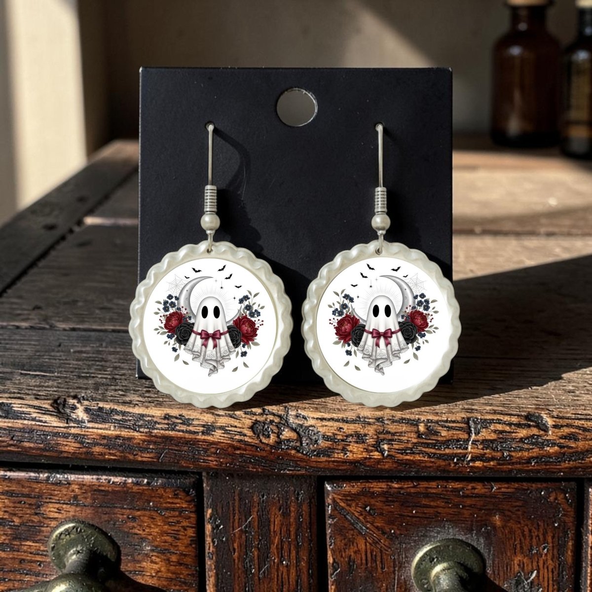 A pair of Crescent Moon Lace Ghost Scalloped Earrings by Mirabilia Boutique, featuring a vintage-inspired ghost face with red roses, are displayed on a black card atop a rustic wooden surface, with sunlight casting delicate shadows.