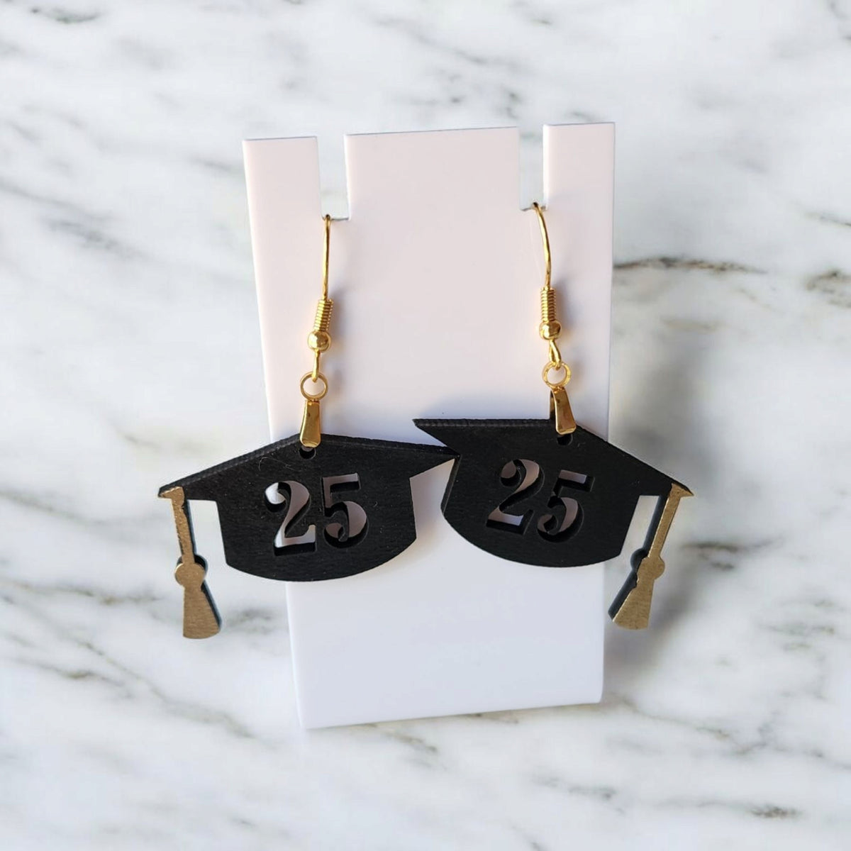 Mirabilia Boutique's Graduation Cap Earrings for the Class of 2025 feature black caps with gold tassels and "25" cutouts—perfect grad gifts or party accessories—shown on a white card against a marble background.