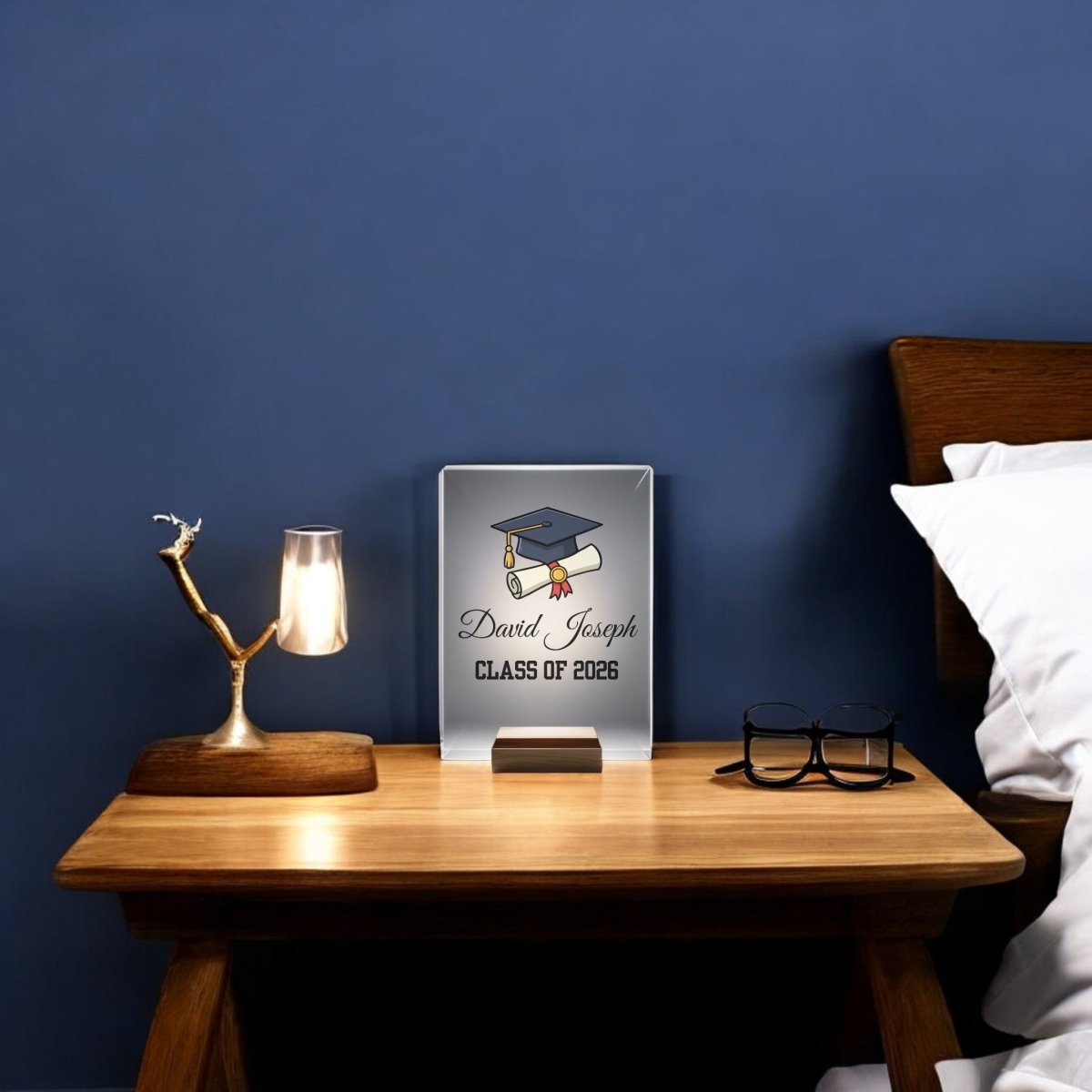 A wooden nightstand with a lamp, tree-shaped jewelry holder, glasses, and a Mirabilia Boutique Classic Graduation Cap Personalized Night Light reading "David Joseph, Class of 2026." The bed and wall appear in the background.
