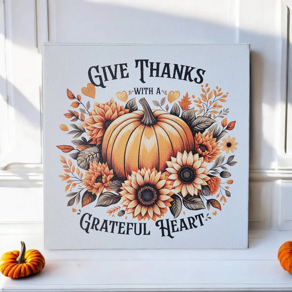 The Mirabilia Boutique Give Thanks With a Grateful Heart 8x8 Wood Sign features pumpkins and sunflowers on cradled wood, perfect as Thanksgiving or fall décor. Two small pumpkins accent the sign on a white surface against a white wall.