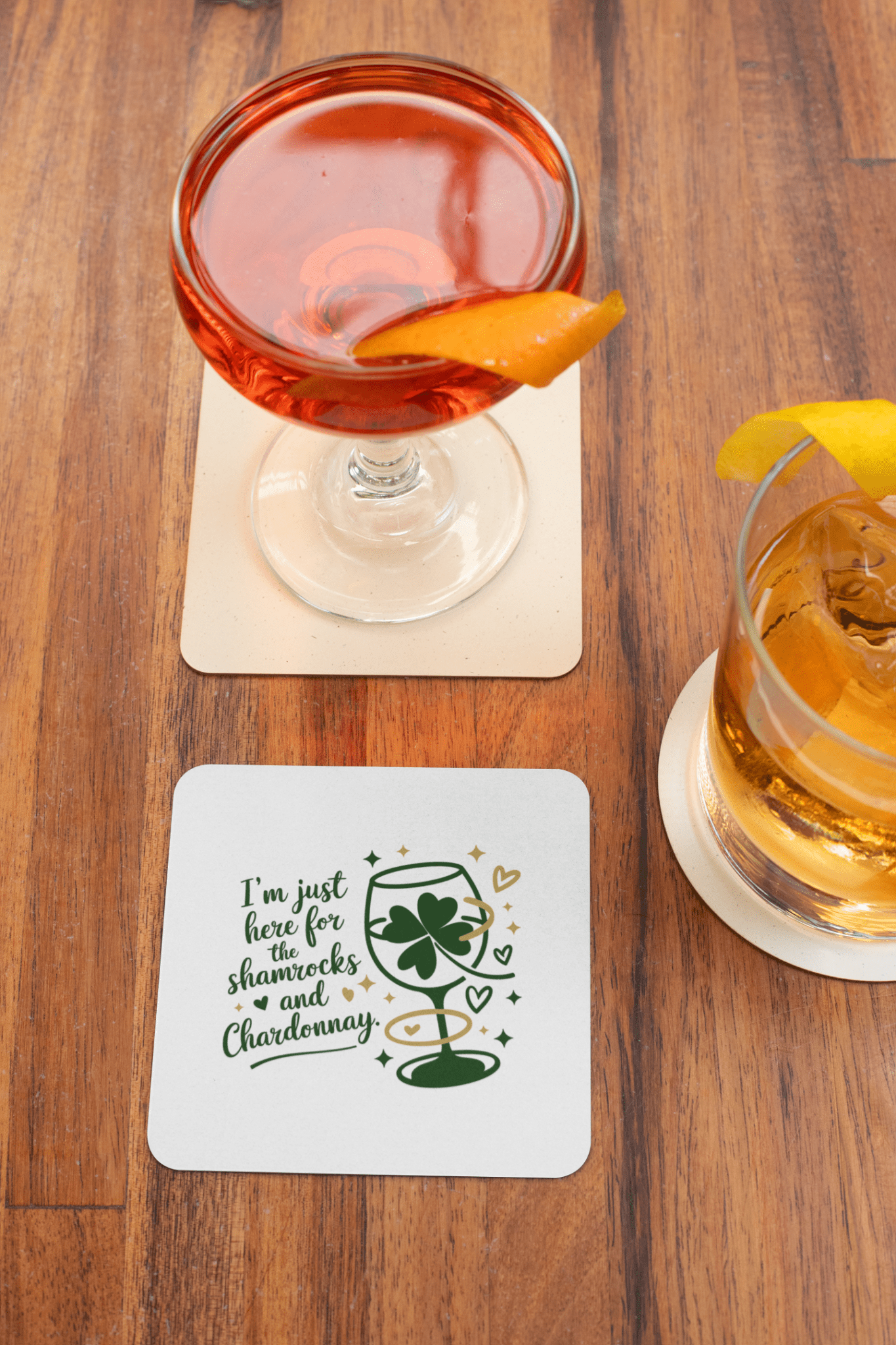 A cocktail and a whiskey glass with orange garnishes sit on coasters atop a wooden table. One is from the Shamrocks and Chardonnay Coaster Set by Mirabilia Boutique, featuring shamrocks, hearts, and “I’m just here for the shenanigans and Chardonnay.”.