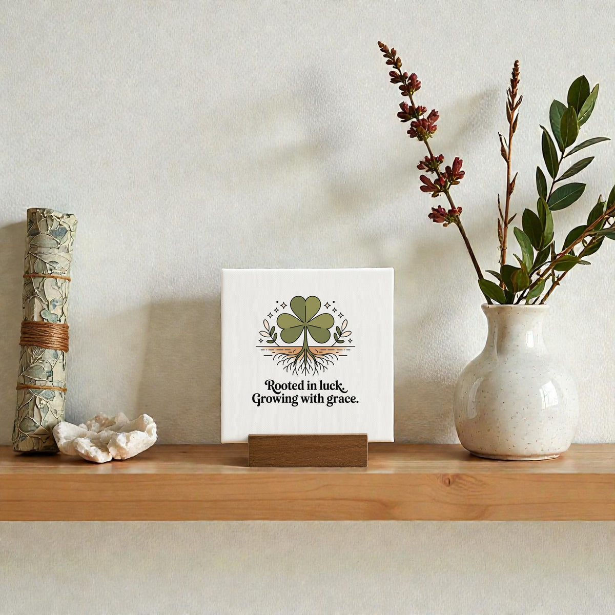 A wooden shelf showcases a scroll, a crystal, mini botanical decor, and the Mirabilia Boutique "Rooted in Luck, Growing with Grace" 4x4 canvas sign featuring a four-leaf clover, plus a ceramic vase with branches bearing small red leaves.