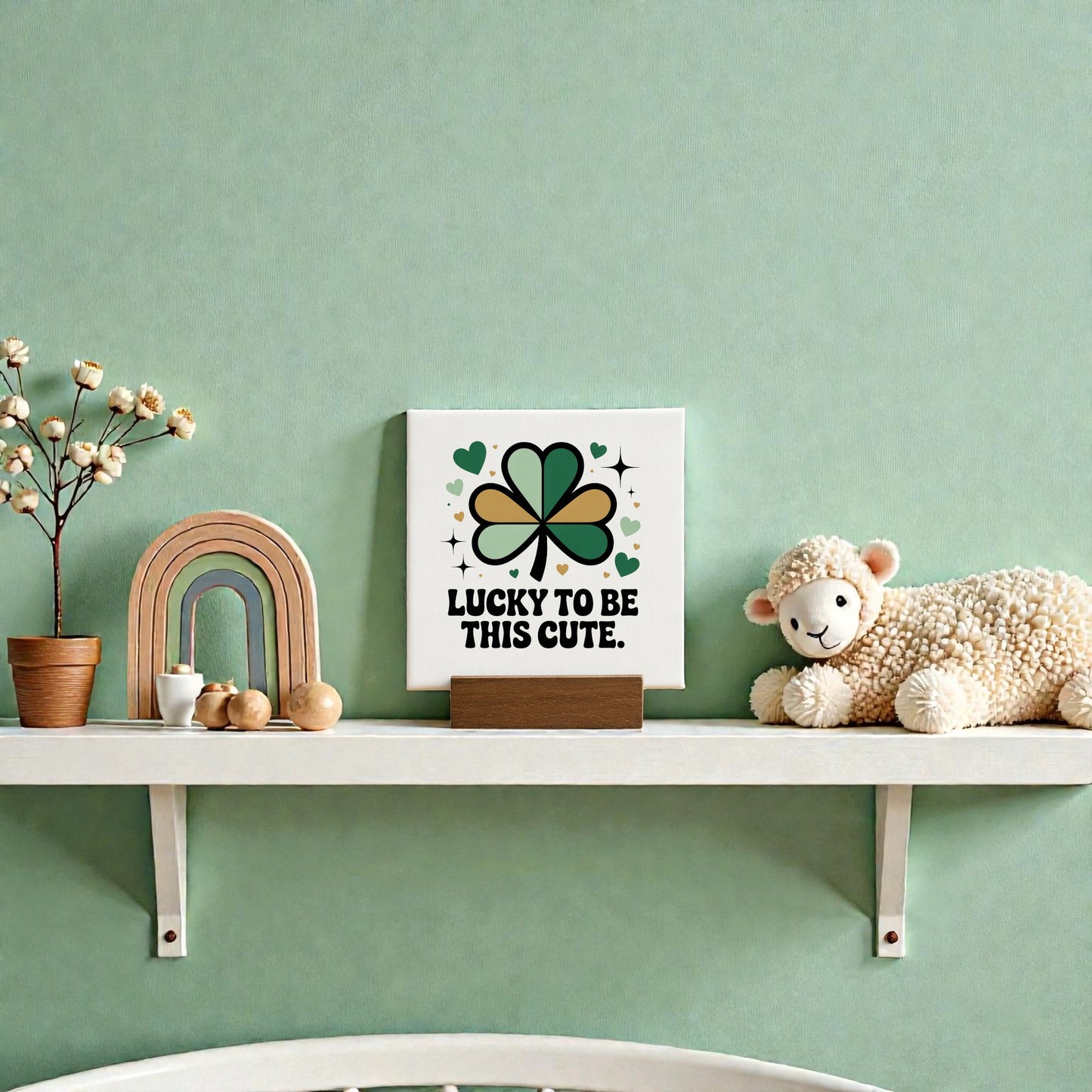 A white shelf displays a Mirabilia Boutique "Lucky to Be This Cute" 4x4 canvas sign, a potted plant, wooden rainbow, clover decor, decorative acorns, and plush sheep—perfect for any little one's St. Patrick’s Day room décor.