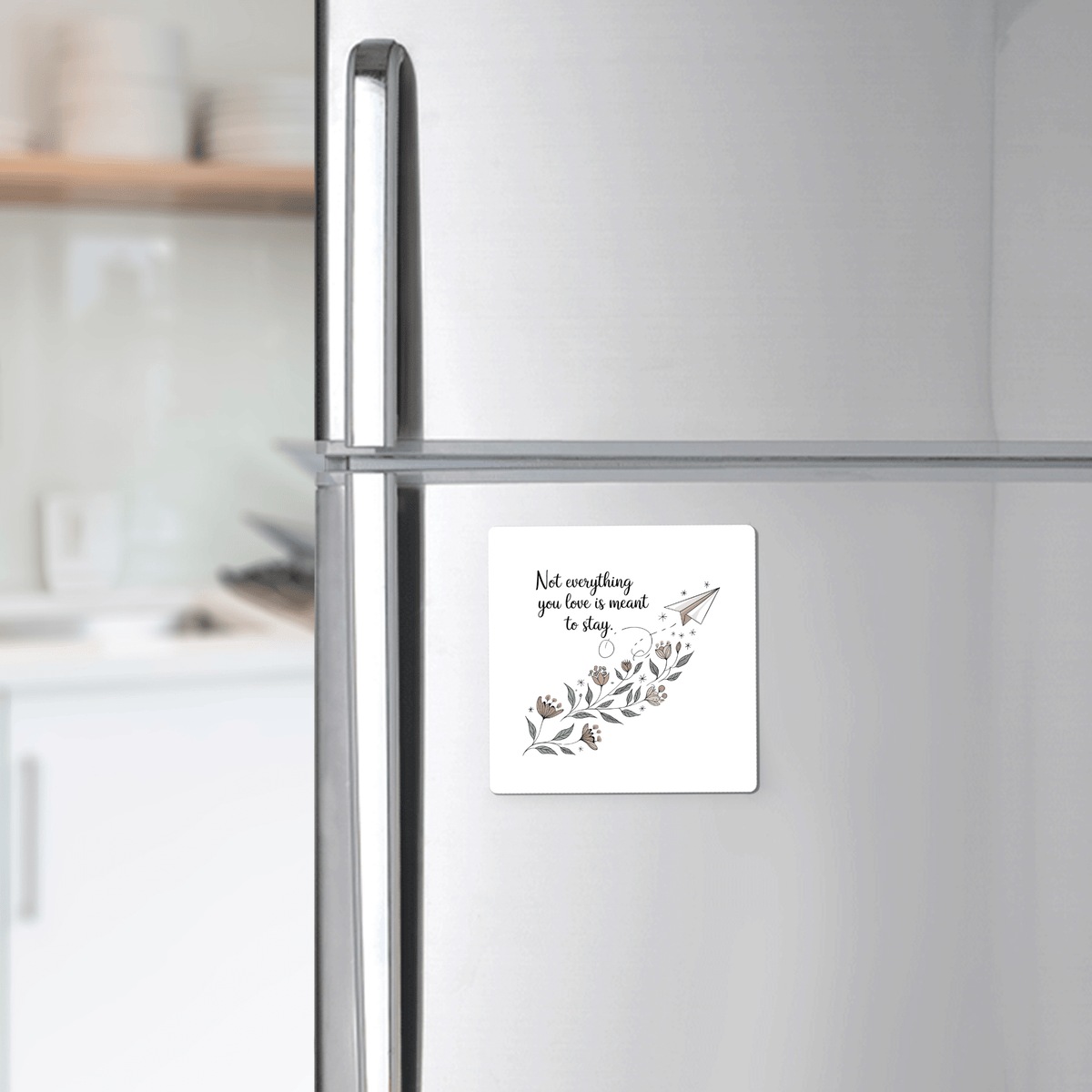 A silver fridge displays the Mirabilia Boutique Grief Quote Magnet, reading "Not Everything You Love Is Meant to Stay," with floral art. Blurred kitchen shelves with dishes are seen in the background.