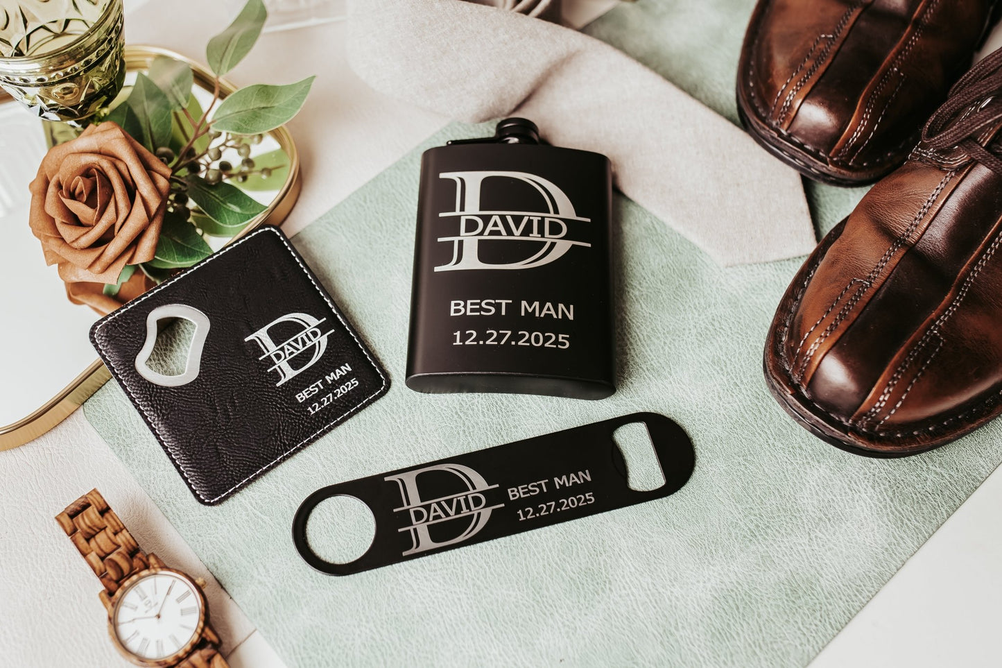 A flat lay features brown dress shoes, a watch, boutonnière, and the Mirabilia Boutique Personalized Best Man Flask Gift Set—engraved “David, Best Man, 12.27.2025”—with a custom flask, bottle opener, and leather keychain on green fabric.