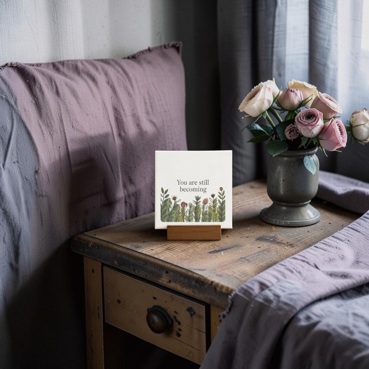 A rustic wooden stand displays the Mirabilia Boutique "You Are Still Becoming" Mini Canvas Sign with botanical art, set beside a vase of pink and white roses near a neatly made bed with lavender bedding.