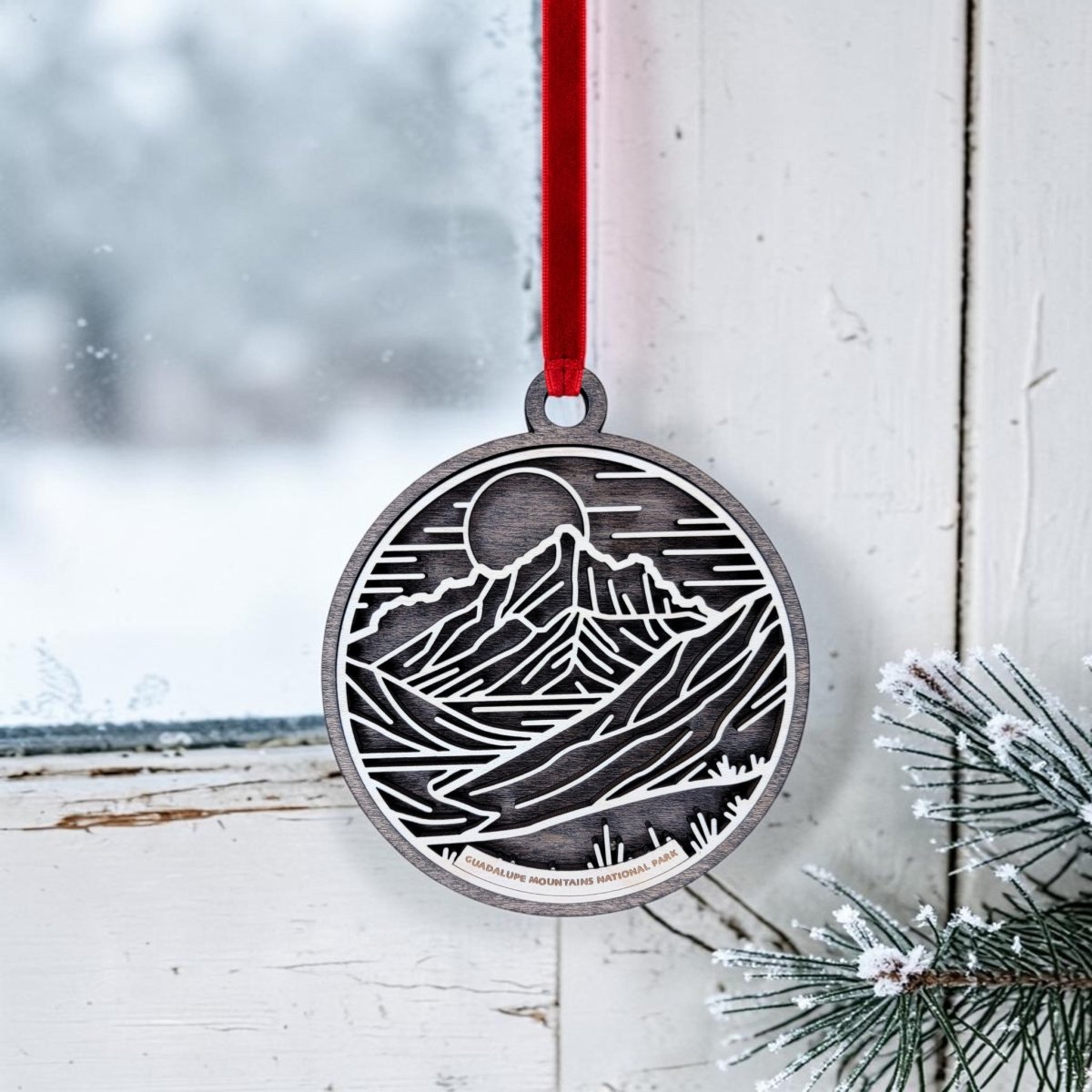 The Guadalupe Mountains National Park Line Art Christmas Ornament by Mirabilia Boutique, featuring a metallic mountain and sun design, hangs from a red ribbon on a white window. Outside, snowy scenery can be seen, while frosted pine branches rest at the windowsill, capturing the peaceful essence of the park.