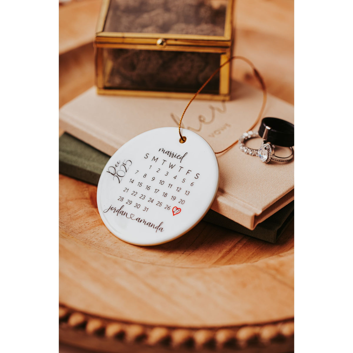 The Personalized Wedding Date Ornament by Mirabilia Boutique is a custom ceramic calendar keepsake featuring a heart around November 25th and "jordan & amanda" below—perfect for newlyweds, displayed beside rings and a jewelry box on books.