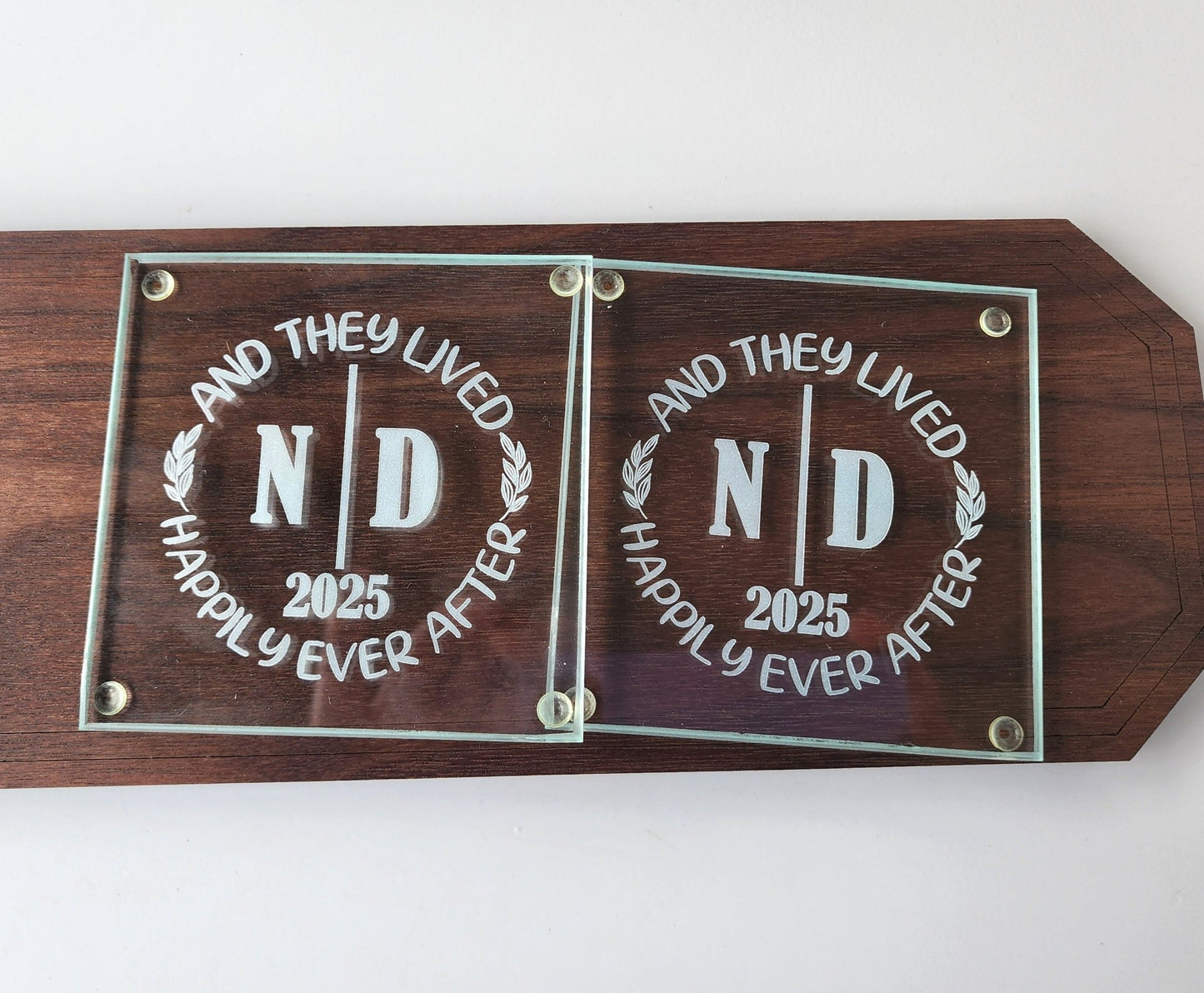 The Personalized Glass Wedding Coasters by Mirabilia Boutique feature elegantly engraved "ND" and "2025" on a wooden plaque with the phrase "And they lived happily ever after," making them an ideal romantic home décor and a perfect personalized wedding gift.