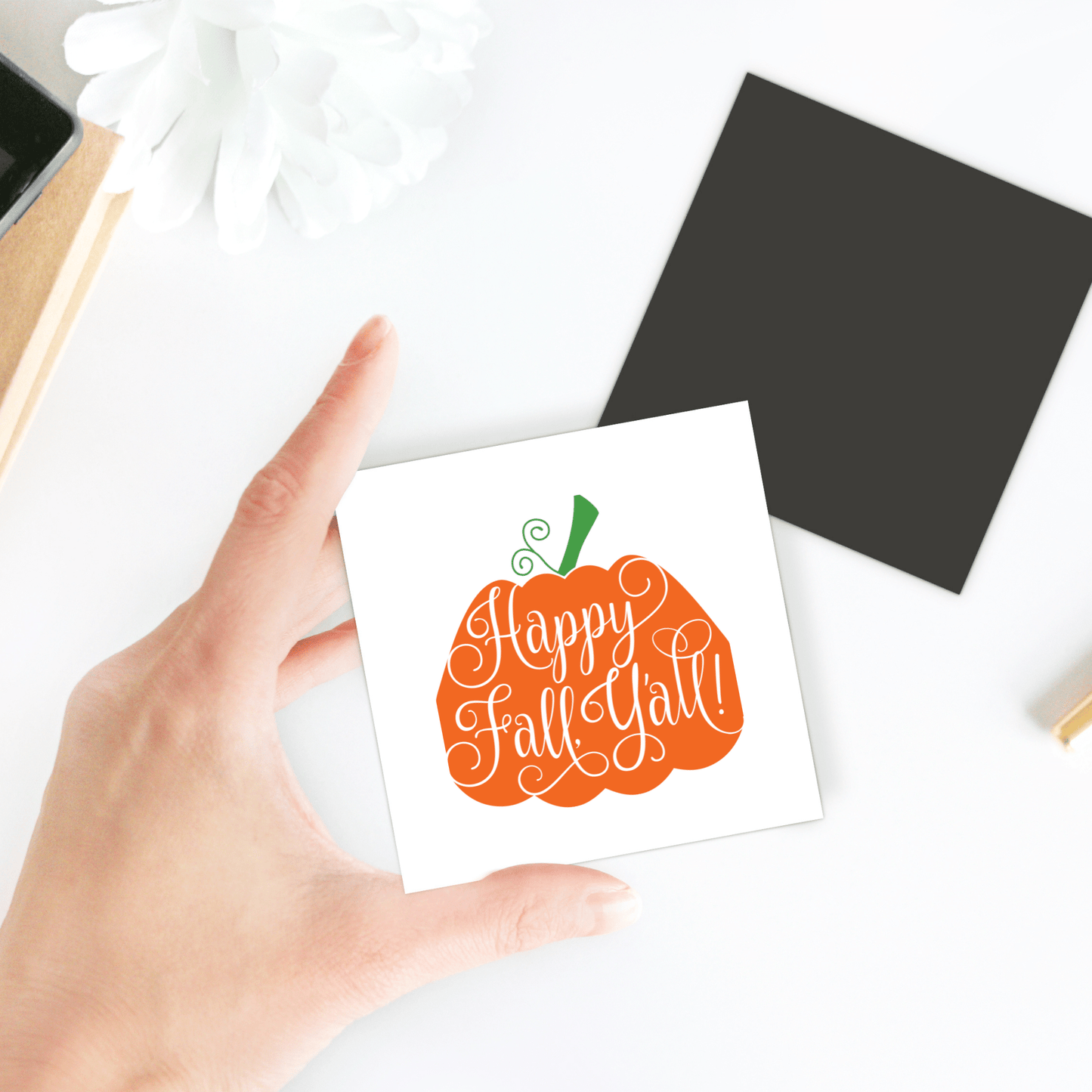 A hand holds Mirabilia Boutique’s "Happy Fall Y’all Magnet"—a pumpkin-shaped fall decor piece with handlettered script. Perfect for your fridge, this festive accent adds autumn charm to any space. Black and white accents appear on the table behind.