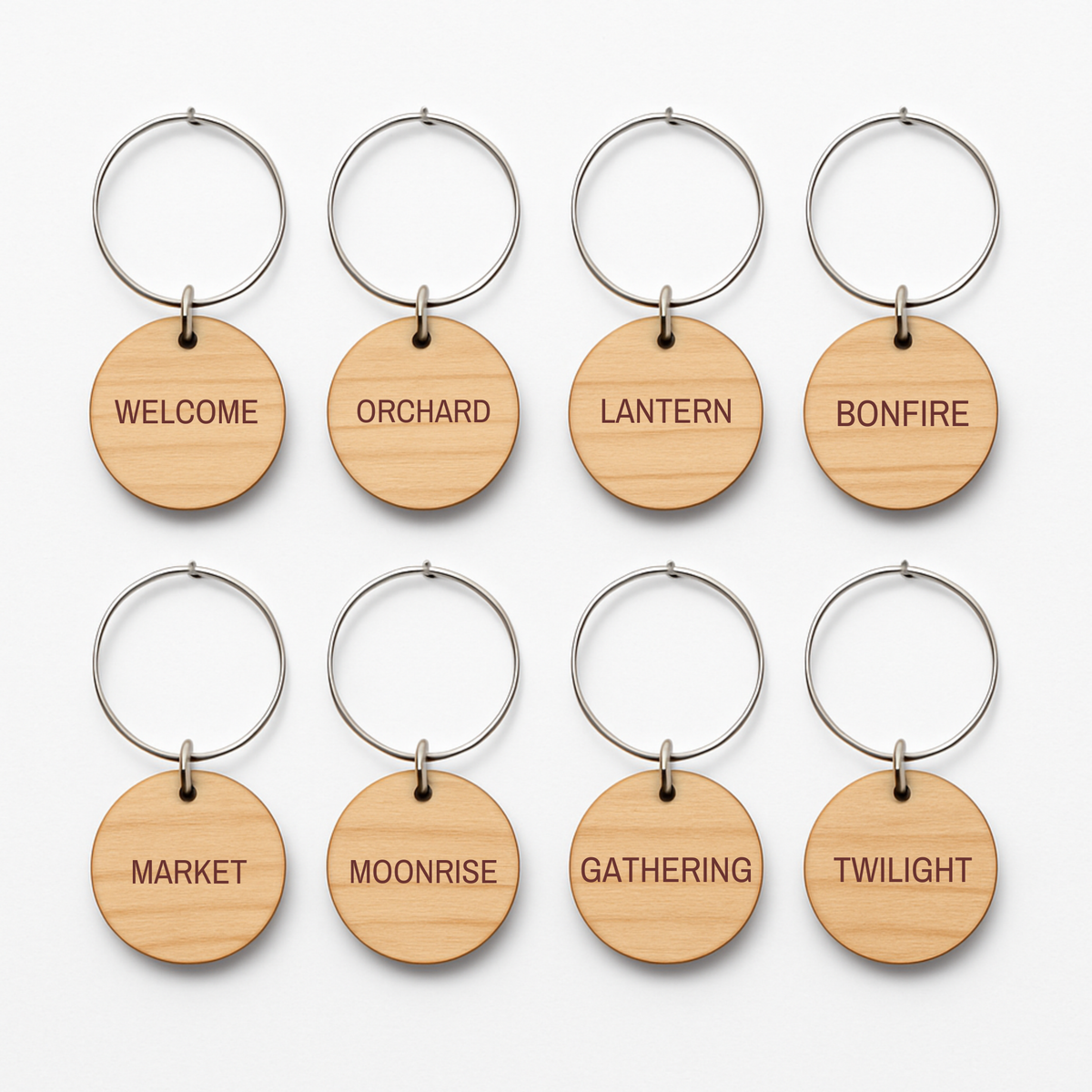 Set of eight engraved wooden charms—WELCOME, ORCHARD, LANTERN, BONFIRE, MARKET, MOONRISE, GATHERING, and TWILIGHT—from Mirabilia Boutique’s Haunted Harvest Rustic Wine Charms Set. Displayed in two rows on a white background; ideal as wine charms or a unique host gift for autumn gatherings.