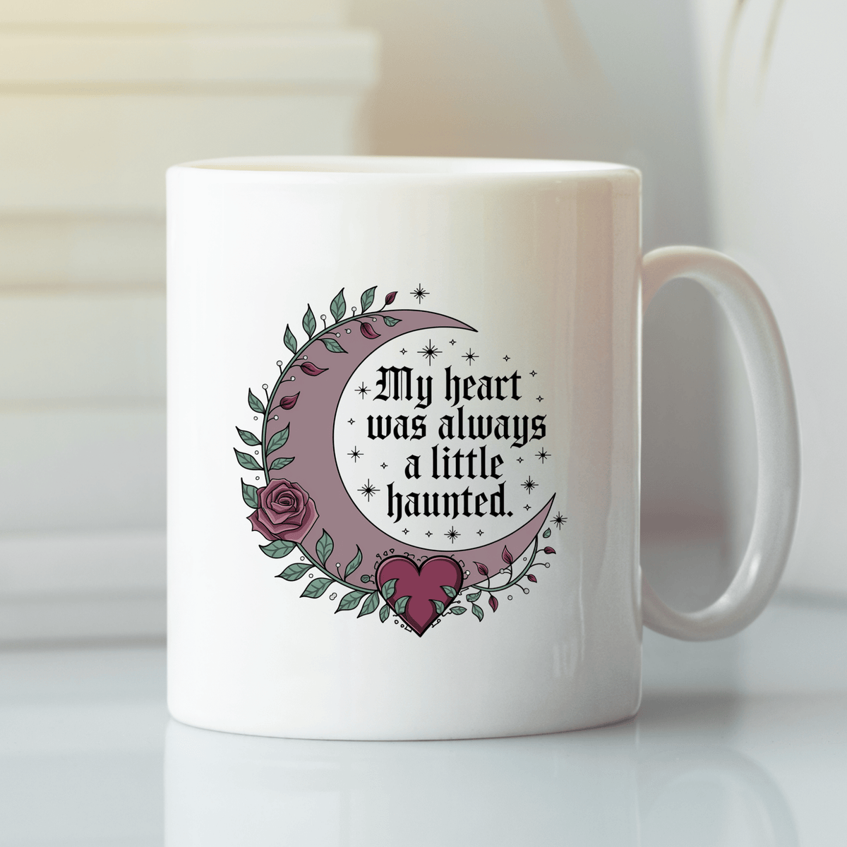 The Mirabilia Boutique Gothic Mug features white ceramic, a crescent moon, leaves, a rose, and a heart with "My Heart Was Always a Little Haunted" in gothic font.