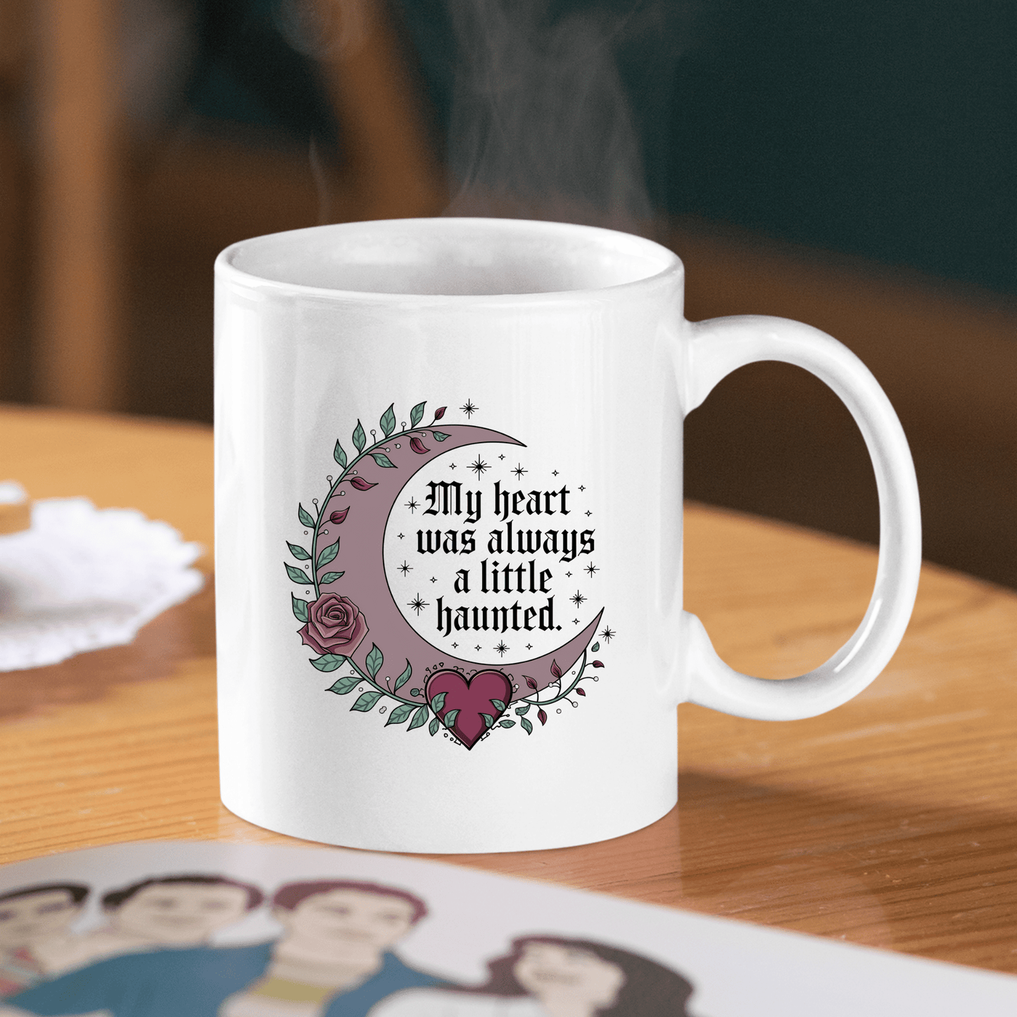 The Gothic Mug | My Heart Was Always a Little Haunted by Mirabilia Boutique features a white design with a crescent moon, rose, and heart, plus the phrase "My heart was always a little haunted." Steam rises as it rests on a wooden table.