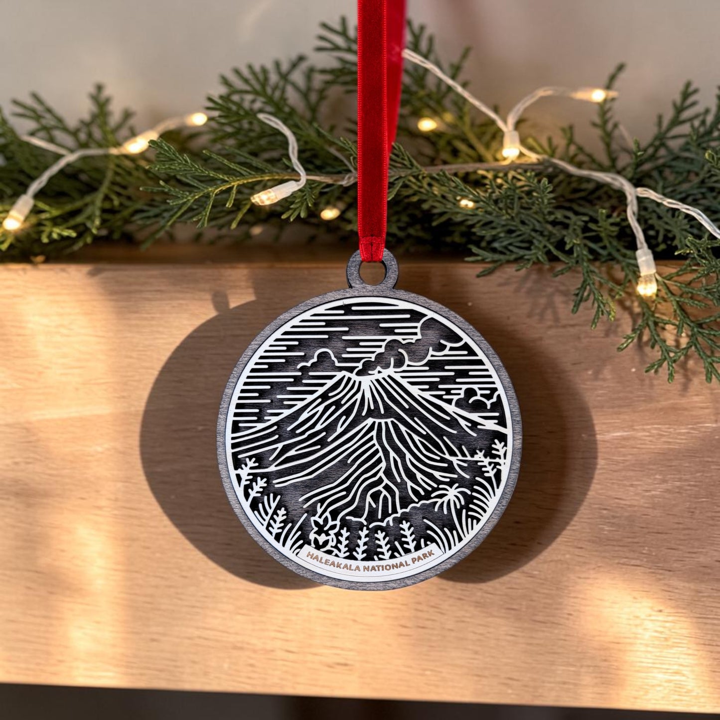 The Haleakalā National Park Line Art Christmas Ornament from Mirabilia Boutique features an oval wooden design with an engraved mountain scene and "Haleakalā National Park" text, hung from a red ribbon—making it a perfect Maui keepsake. Pine branches and string lights add festive charm to the background.