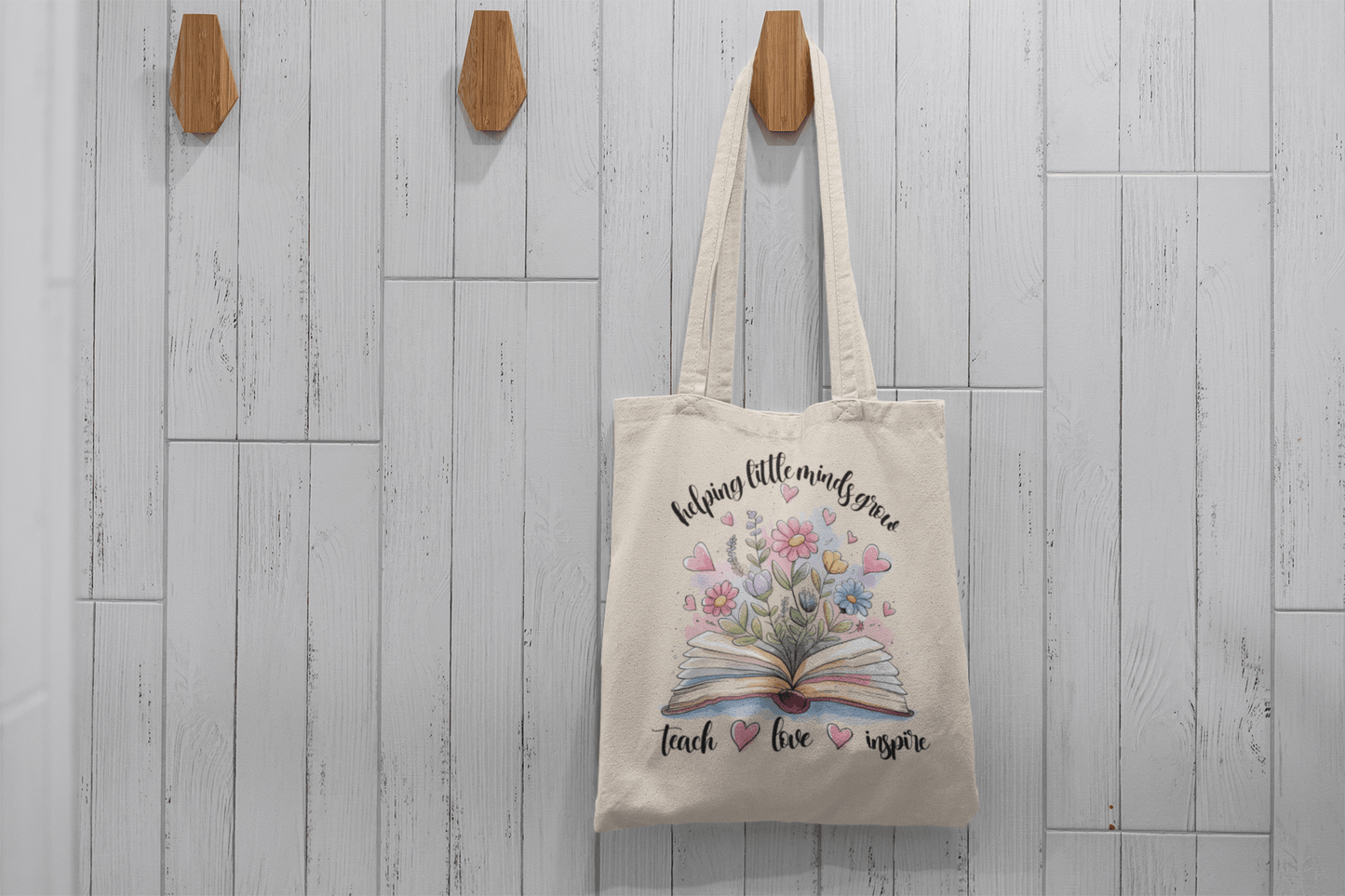 The Helping Little Minds Grow Tote Bag by Mirabilia Boutique hangs on a hook against a white tiled wall. This beige canvas teacher bag features flowers, an open book, pink hearts, and the words "Helping little minds grow"—a perfect gift.
