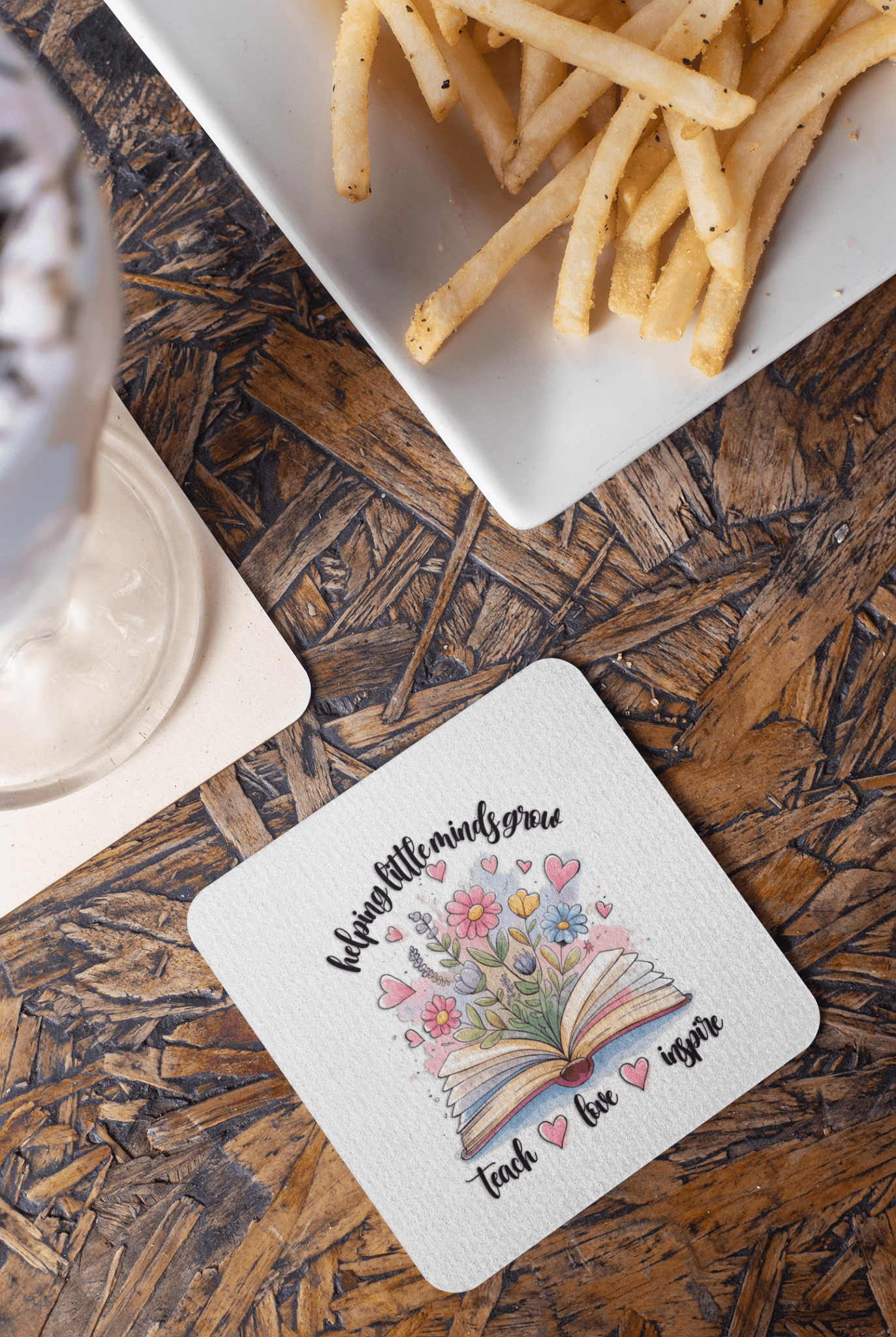 The Mirabilia Boutique Helping Little Minds Grow Coasters (Set of 4) feature an open book with flowers, hearts, and the message "Helping little minds grow, Teach ♥ Love ♥ Inspire"—a perfect teacher gift.