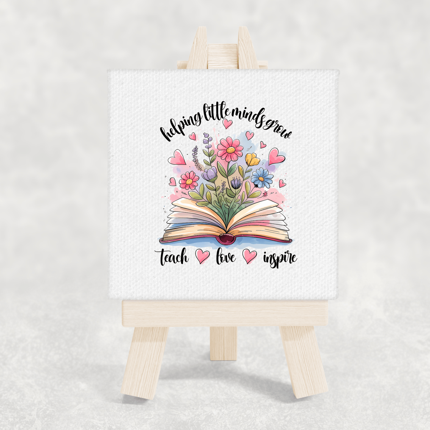 The Mirabilia Boutique Helping Little Minds Grow Mini Canvas with easel displays an open book with flowers and hearts, plus the phrase “helping little minds grow, teach, love, inspire”—a perfect teacher gift or classroom decor.