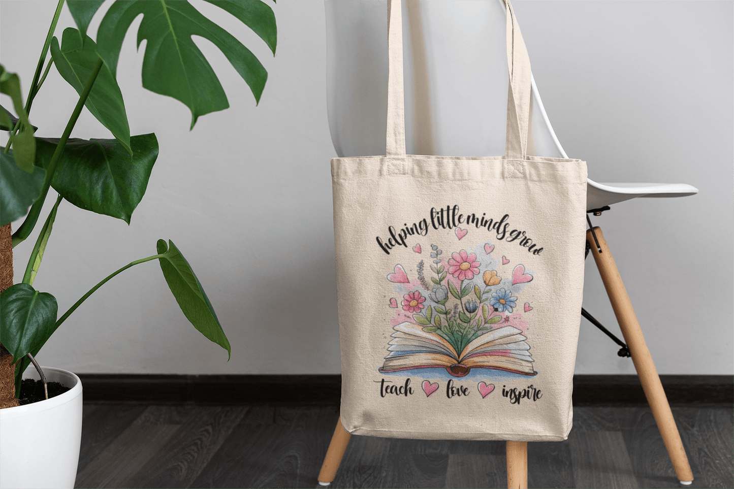 The Mirabilia Boutique "Helping Little Minds Grow" Teacher Canvas Tote Bag features an open book with blooming flowers and the phrase "Teach. Love. Inspire."—a perfect teacher appreciation gift, pictured beside a green potted plant.