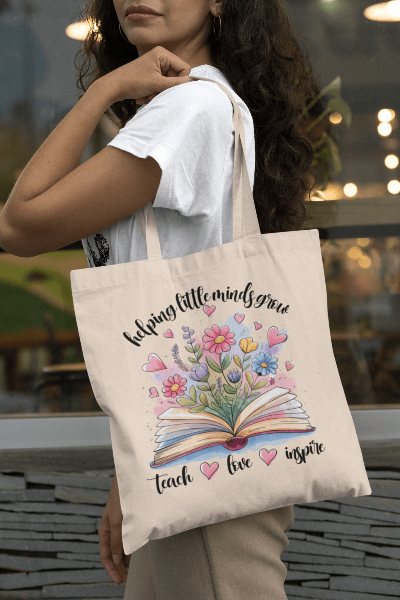 A woman holds the Mirabilia Boutique "Helping Little Minds Grow Tote Bag," a teacher canvas bag featuring an open book with flowers and hearts, and the phrase: "helping little minds grow, teach, love, inspire.