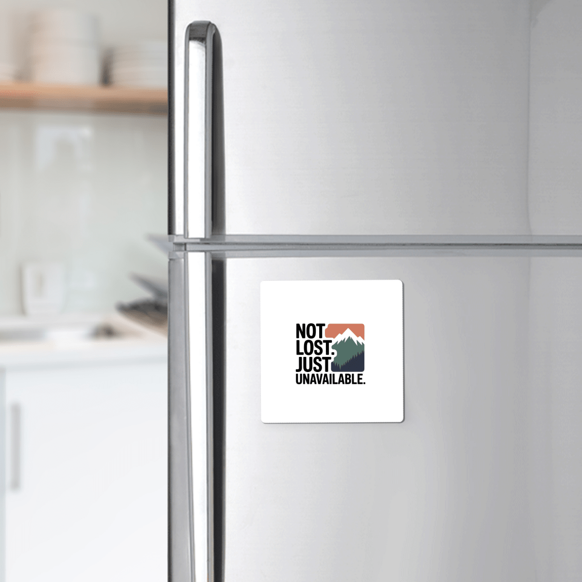 The Not Lost Just Unavailable Adventure Magnet by Mirabilia Boutique features a mountain graphic and the phrase "NOT LOST. JUST UNAVAILABLE."—a unique outdoor gift for adventurers, shown on a stainless steel fridge with a blurred kitchen in the background.