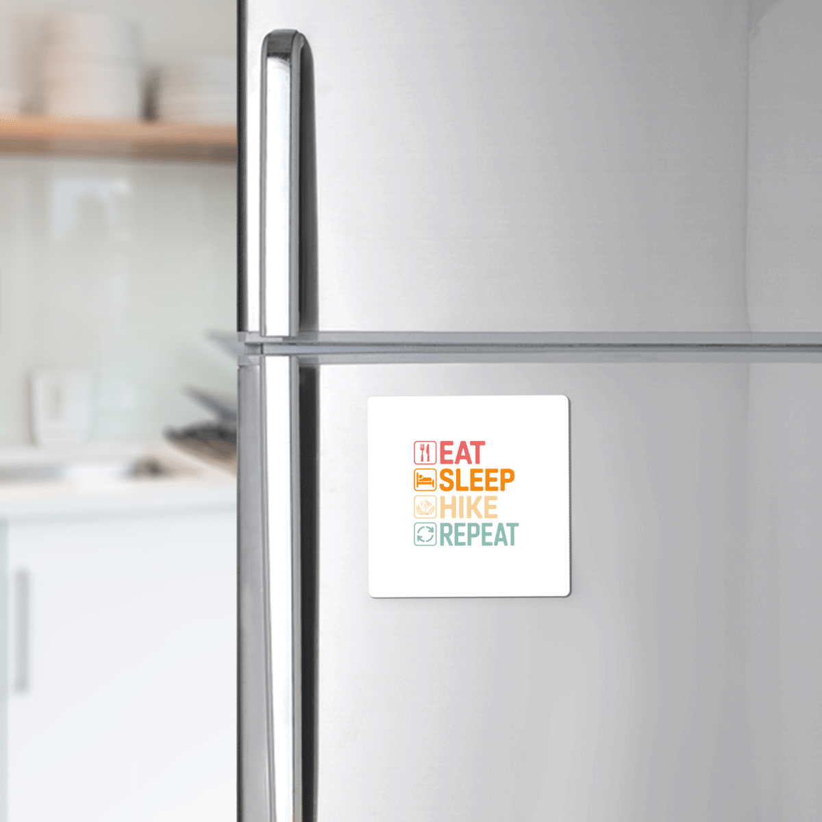 A stainless steel refrigerator features the "Eat Sleep Hike Repeat Magnet" by Mirabilia Boutique, with colorful bold letters. The softly blurred kitchen background makes this a perfect hiking gift for outdoor lovers.