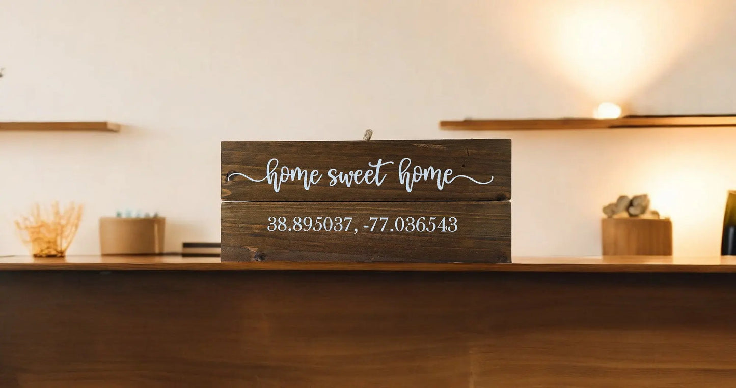 A charming Home Sweet Home Wall Plaque from Mirabilia Boutique rests on the countertop, featuring the customizable coordinates "38.895037, -77.036543." The softly lit room in the backdrop, adorned with shelves and decorative items, enhances the cozy rustic ambiance.