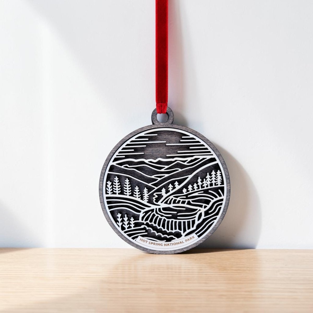 The Hot Springs National Park Line Art Christmas Ornament by Mirabilia Boutique is a handcrafted wooden round medal, adorned with a black and white landscape illustration of mountains, trees, and a winding path. It hangs from a red ribbon against a white wall and wooden surface, beautifully capturing the essence of Hot Springs National Park.