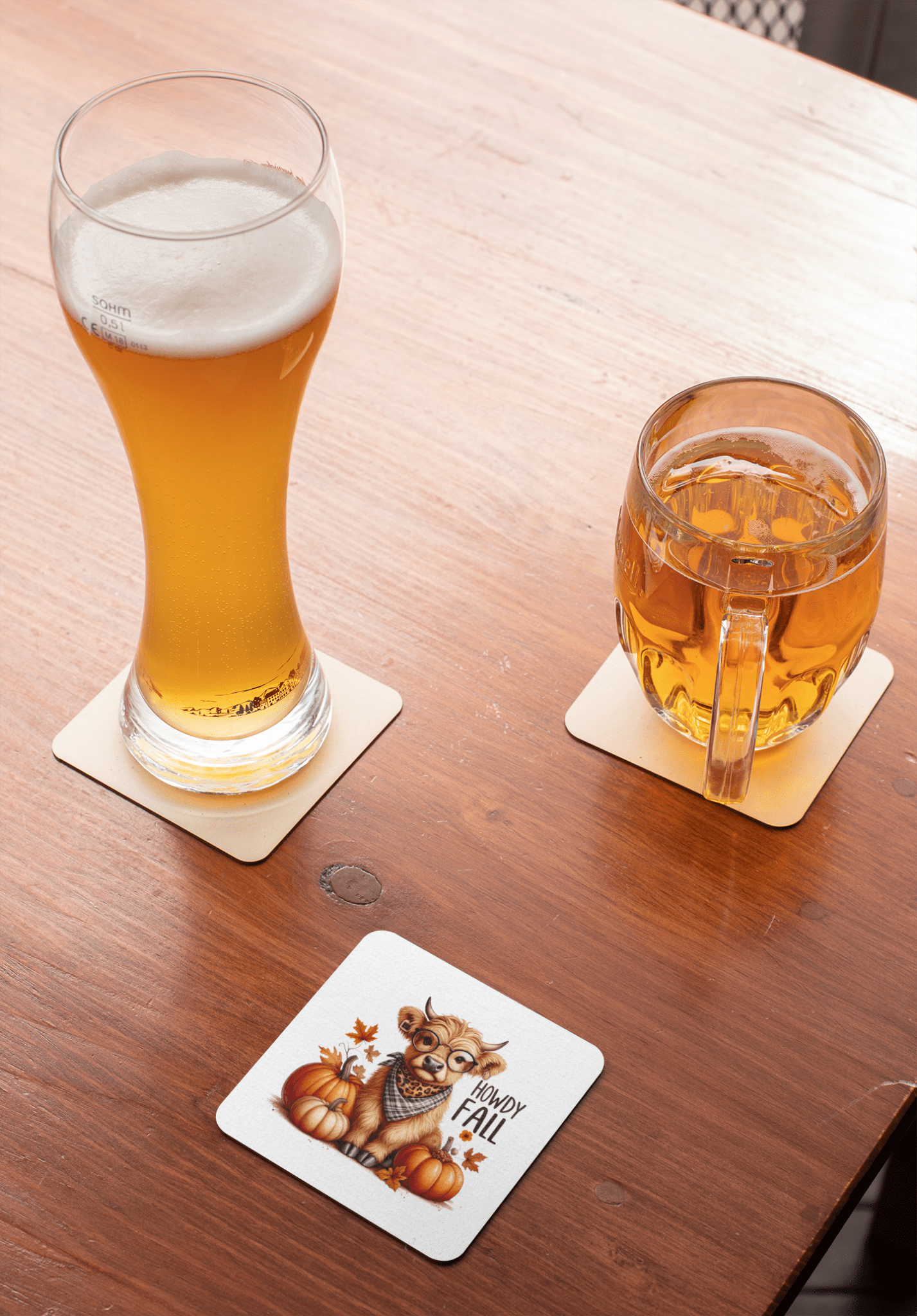 A beer and amber drink sit on coasters atop a wooden table. A third Howdy Fall Highland Cow coaster by Mirabilia Boutique features a cow with glasses and pumpkins, reading "Happy Fall!"—a cute western autumn decor accent.