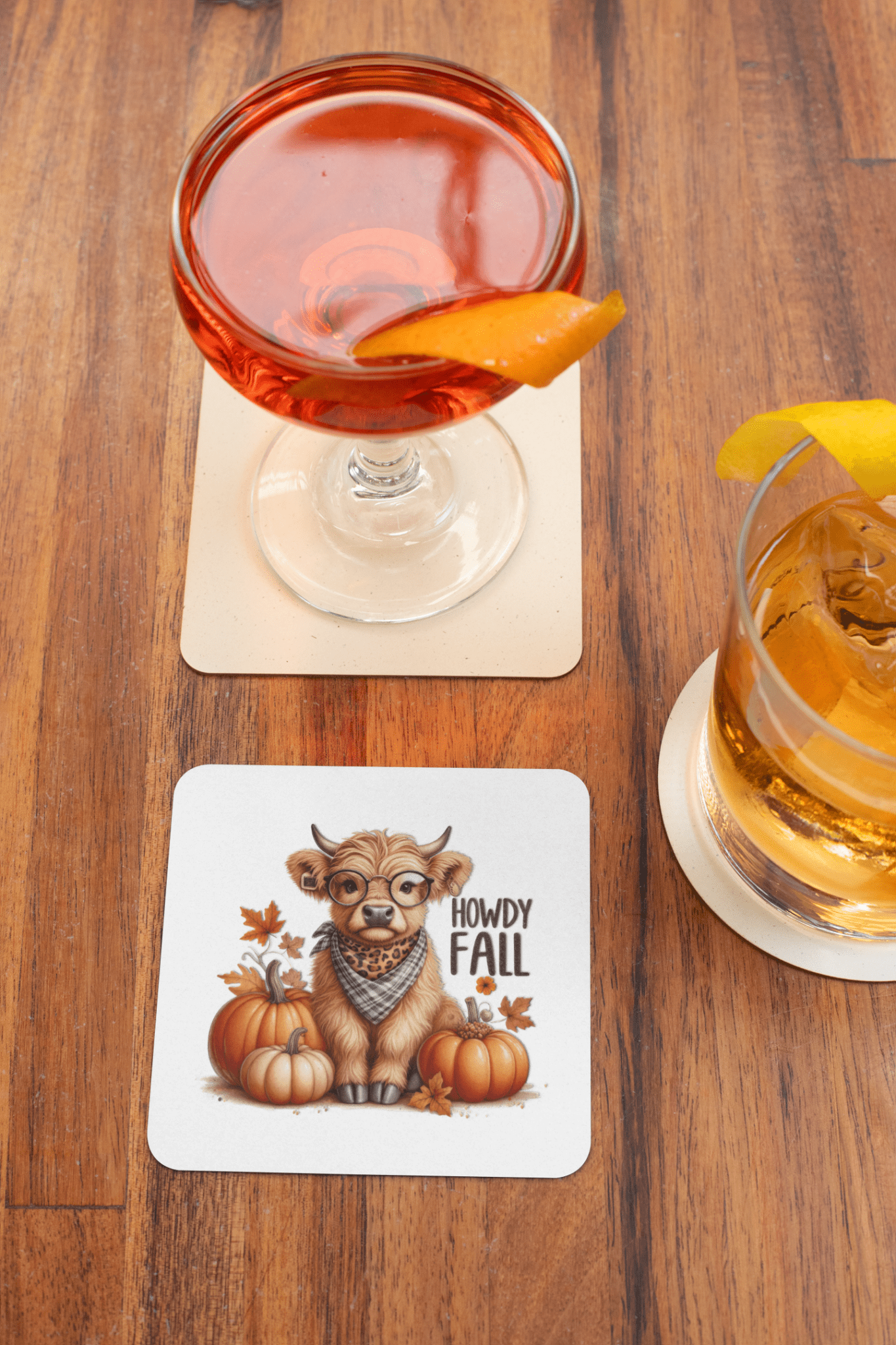 A red cocktail and a brown drink with citrus garnishes sit on coasters. A third coaster displays Mirabilia Boutique’s "Howdy Fall Highland Cow" design—an autumn cow in glasses and scarf, with pumpkins, leaves, and fall decor.