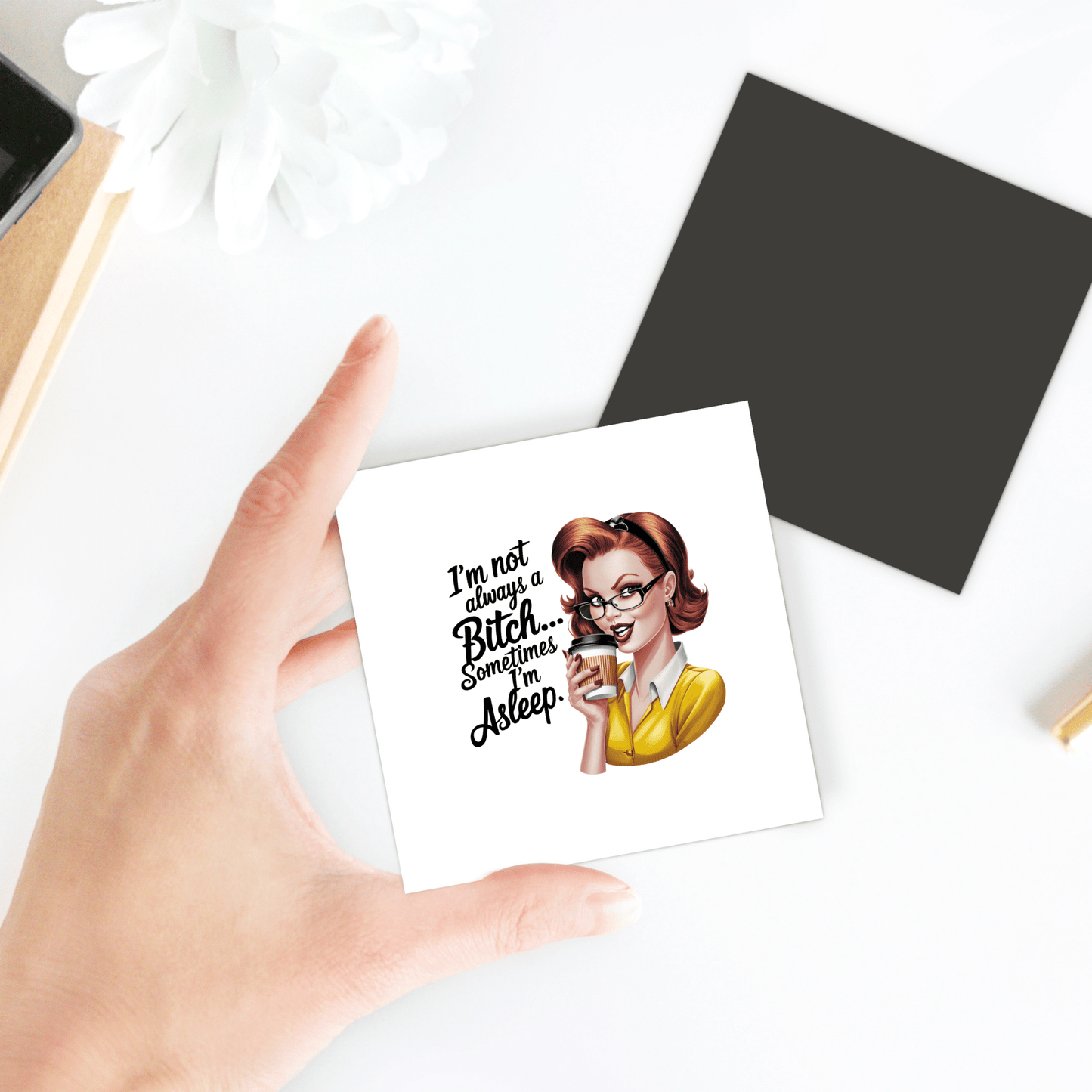A hand holds the "I'm Not Always a Bitch" magnet by Mirabilia Boutique—a funny 3x3 retro-style UV printed magnet, perfect as a coffee and sass gift for friends or coworkers. The magnet rests near a black square and white flower on the table.