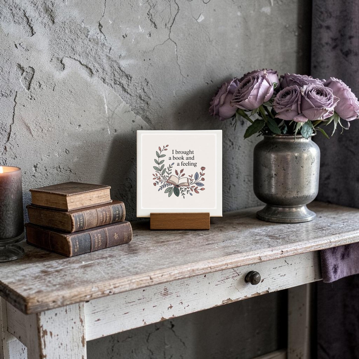 A rustic table displays vintage books, a lit candle, purple flowers in a vase, and the Mirabilia Boutique "I Brought a Book and a Feeling" mini sign—perfect reading decor for any book lover’s gift.