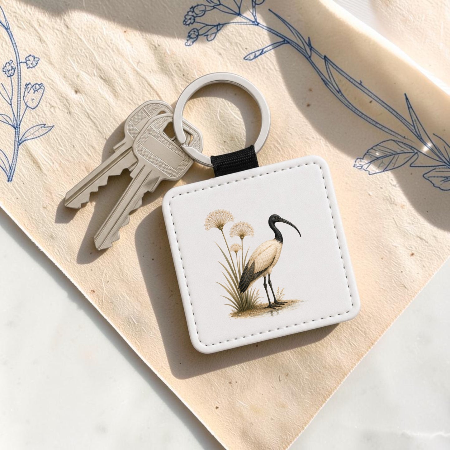 The Ibis & Papyrus Keychain | Ancient Egyptian Art by Mirabilia Boutique is a square keychain adorned with an illustrated white ibis and papyrus standing amid grass. It is shown attached to two silver keys, resting on a sheet of paper decorated with blue floral line art reminiscent of ancient Egyptian motifs.