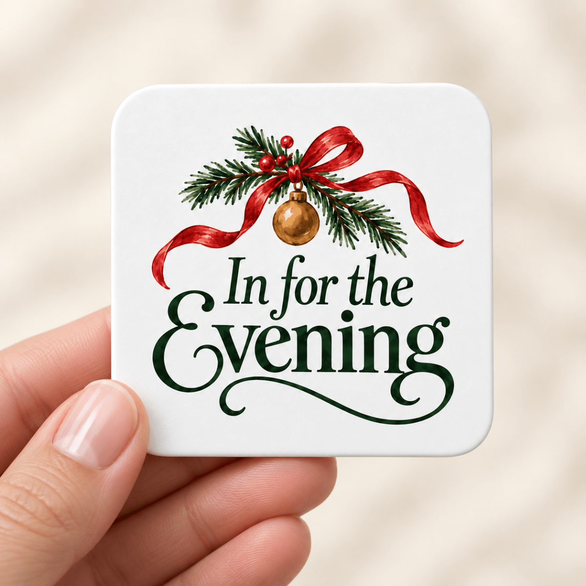 A hand holds a square In for the Evening Christmas Magnet | Mirabilia decorated with pine branches, a gold ornament, a red ribbon, and the text “In for the Evening.”