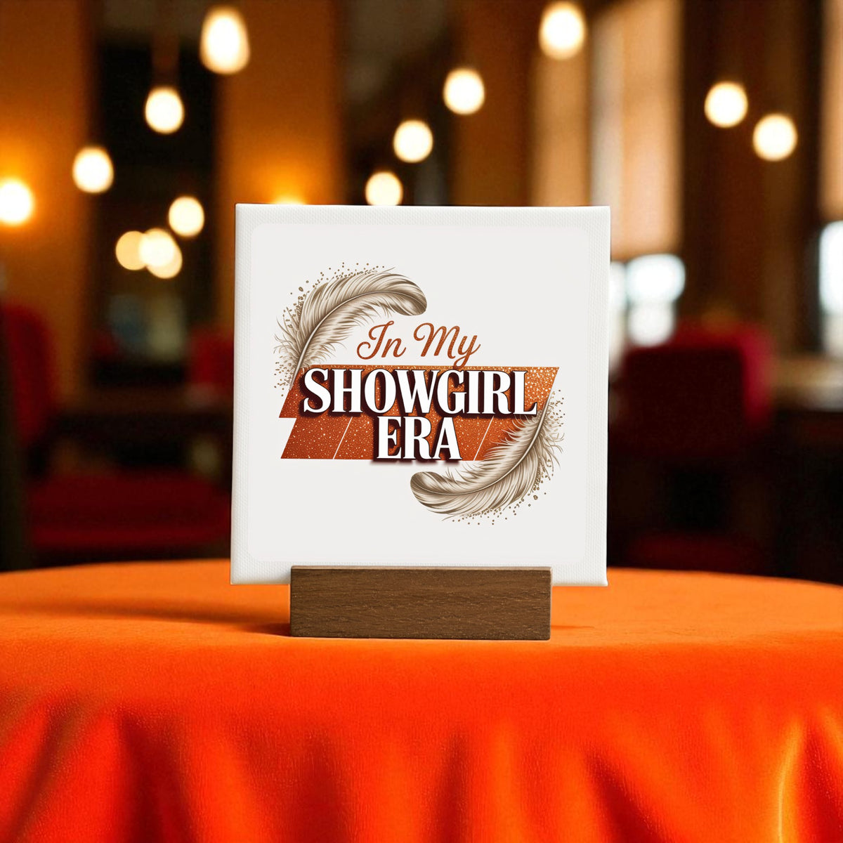 The "In My Showgirl Era Mini Sign" by Mirabilia Boutique adds retro glam to an orange table, featuring bold lettering and feathers, in a warmly lit room with blurred lights and red chairs in the background.