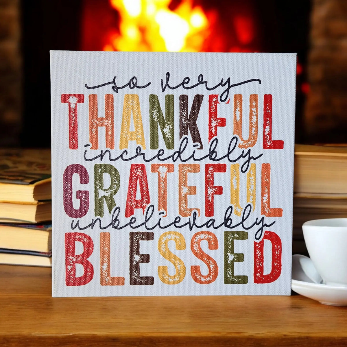 A Thankful Grateful Blessed Inspirational Sign from Mirabilia Boutique, featuring the heartfelt message "So very thankful, incredibly grateful, unbelievably blessed," is displayed against a cozy background with a fireplace, stacks of books, and a white coffee cup and saucer. The vibrant Thanksgiving colors add warmth to this charming seasonal home decor scene.