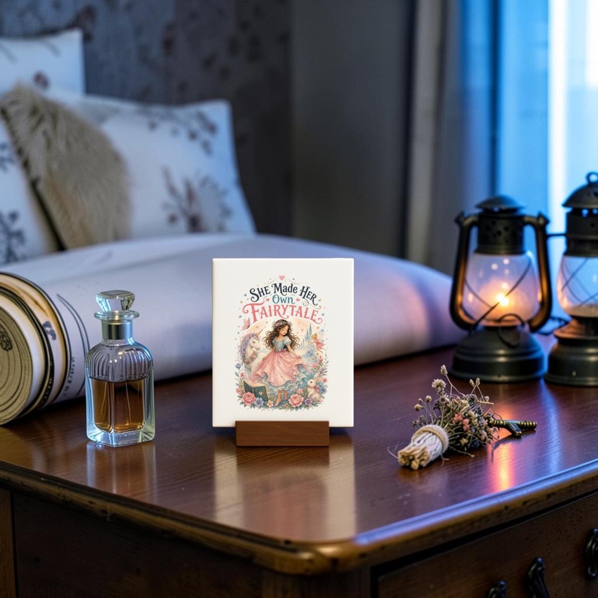 A "She Made Her Own Fairytale Mini Canvas" by Mirabilia Boutique stands on a wooden desk beside a perfume bottle, dried flowers, two lanterns, and a rolled paper. Perfect as unicorn wall art or whimsical children’s room decor, this charming scene features a softly lit bed with pillows in the background.