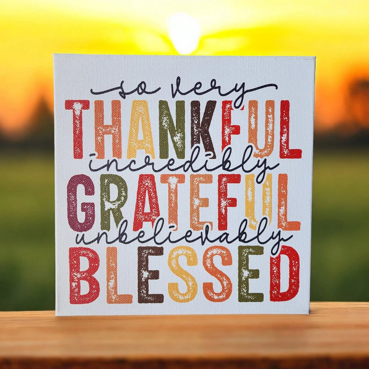 The Mirabilia Boutique "Thankful Grateful Blessed" 8x8 wood sign features an autumn quote on canvas, making it perfect Thanksgiving or Fall décor for your wall or tiered tray. A blurred sunset over a green field adds warmth to your space.