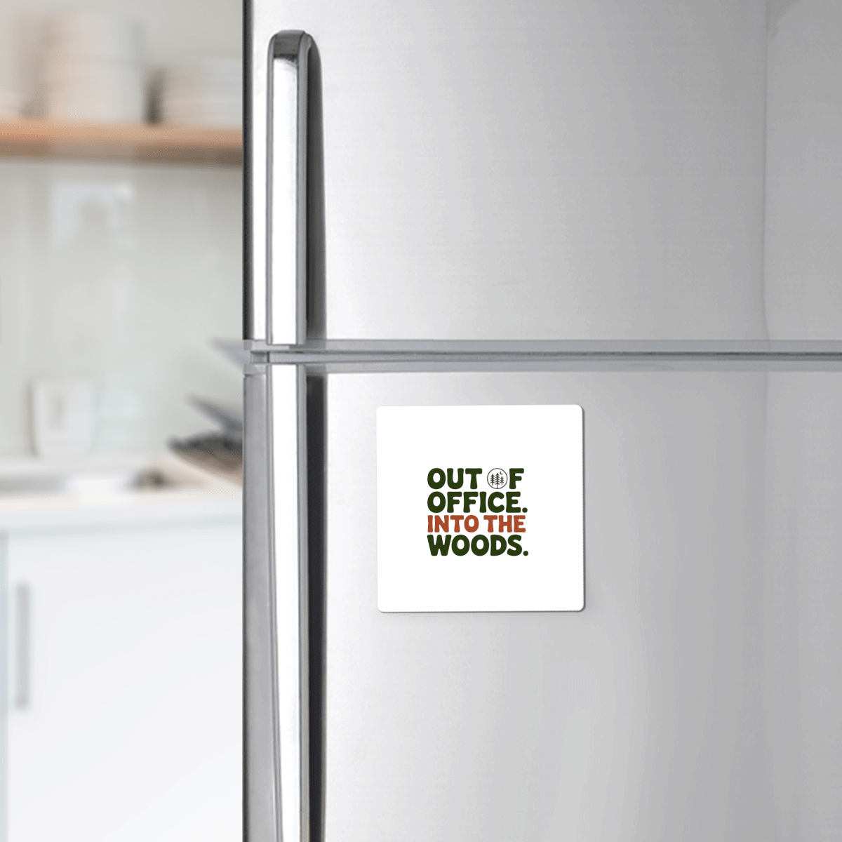 The Out of Office Into the Woods Magnet by Mirabilia Boutique—a square white magnet with green text—is shown on a stainless steel fridge, perfect as cabin decor or a gift for nature lovers. White dishes appear blurred in the kitchen background.