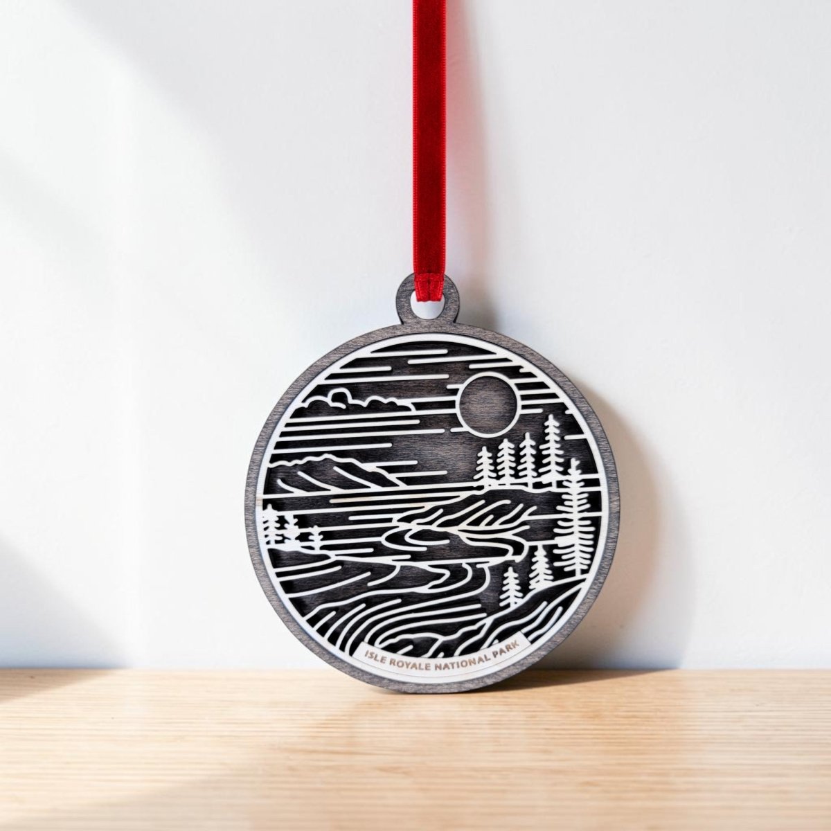 The Isle Royale National Park Line Art Christmas Ornament by Mirabilia Boutique—a round wooden ornament featuring a black and white landscape with trees, water, and the sun inspired by Isle Royale National Park on Lake Superior—hangs from a red ribbon against a white wall, resting on a light wooden surface.