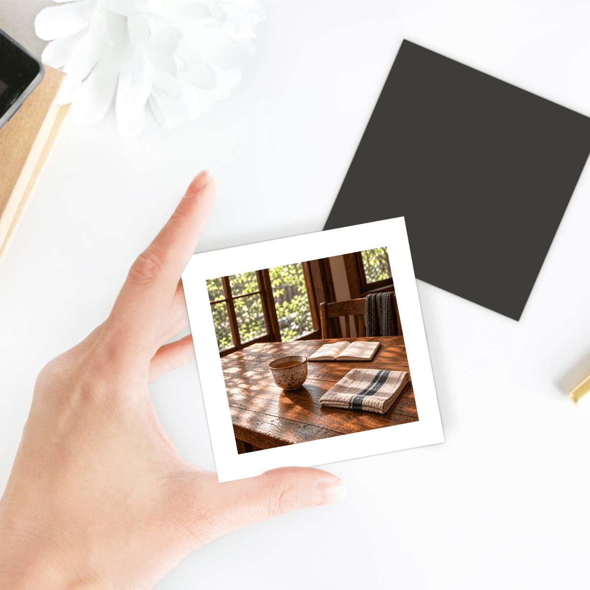 A hand holds The Morning Table Decorative Magnet, a square piece featuring a miniature art scene of a morning setting with a tea cup, an open book, and a folded cloth.