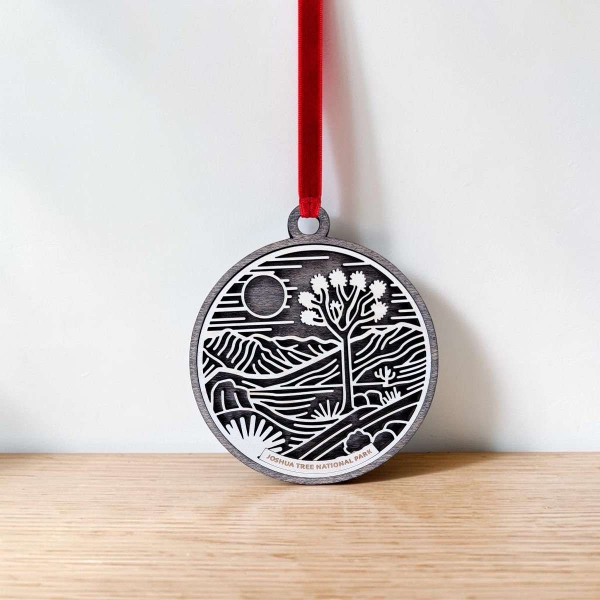 The Joshua Tree National Park Line Art Christmas Ornament by Mirabilia Boutique hangs from a red ribbon against a white wall and wooden surface, showcasing a round wooden design featuring the iconic Joshua tree, sun, and mountains in a desert landscape.