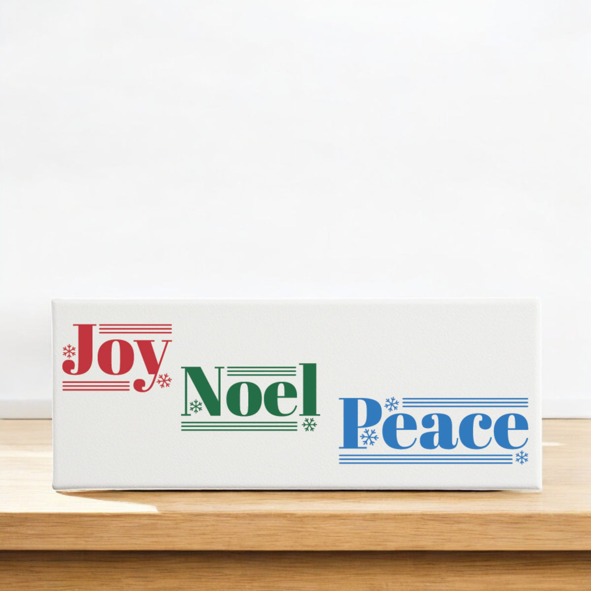 The Mirabilia Boutique Joy Noel Peace Christmas Sign is a white 4x12 canvas shelf sitter featuring "Joy" in red, "Noel" in green, and "Peace" in blue with festive lines and snowflakes—perfect holiday decor for any space.