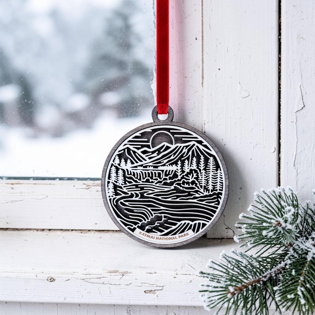 A Katmai National Park Line Art Christmas Ornament by Mirabilia Boutique, showcasing a handcrafted wooden design with a nature-inspired scene and "Katmai National Park" text, hangs from a red ribbon on a snowy window frame. Frosted pine branches in the corner evoke the wild beauty of Alaska brown bears and their pristine habitat.